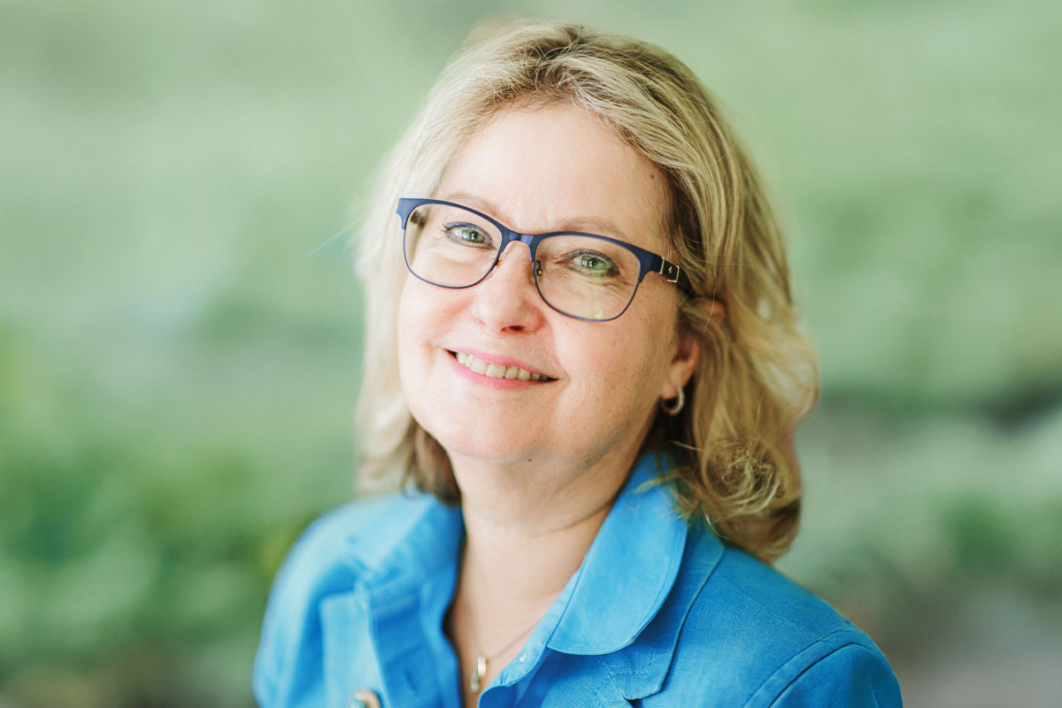 Woman with glasses and medium length blond hair smiles at the camera against a green background