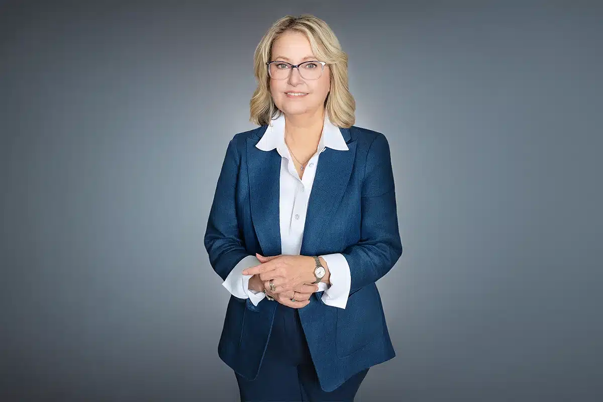 Priscilla's Headshot - blond woman standing with her hands clasped in front of her waist, she's wearing a blue blazer with a white collared shirt underneath